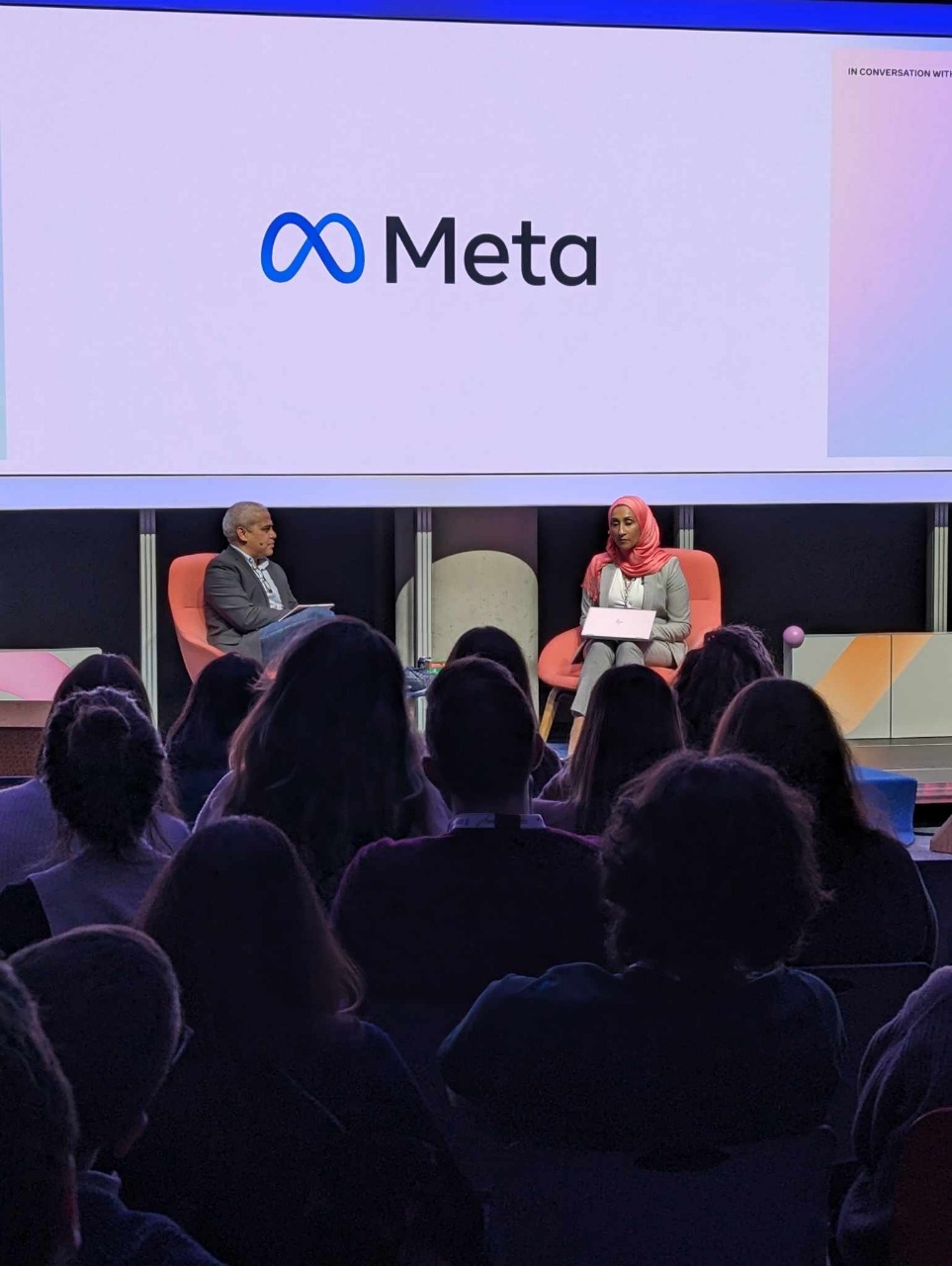 Sam, seated on the right and wearing a coral hijab and light gray outfit, participates in a live panel discussion hosted by Meta. She is holding notes while addressing the audience alongside a male moderator seated to the left. The large Meta logo is displayed prominently on the screen behind them. The venue is filled with attendees seated in rows, attentively listening to the conversation under stage lighting.