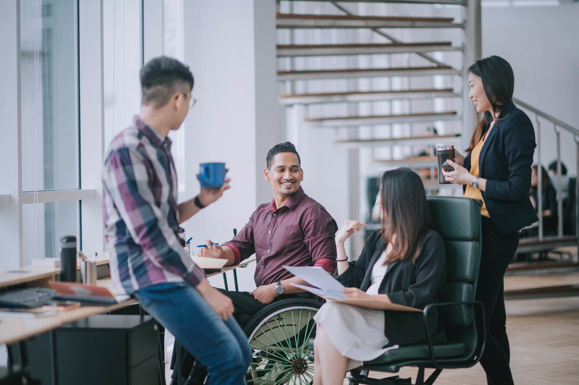 A diverse group of professionals, including a man in a wheelchair, collaborate casually in a modern office with natural light and open stairs