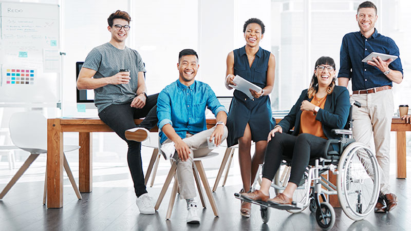A diverse group of professionals, including a woman in a wheelchair, smiling and posing together in a modern, well-lit office space.