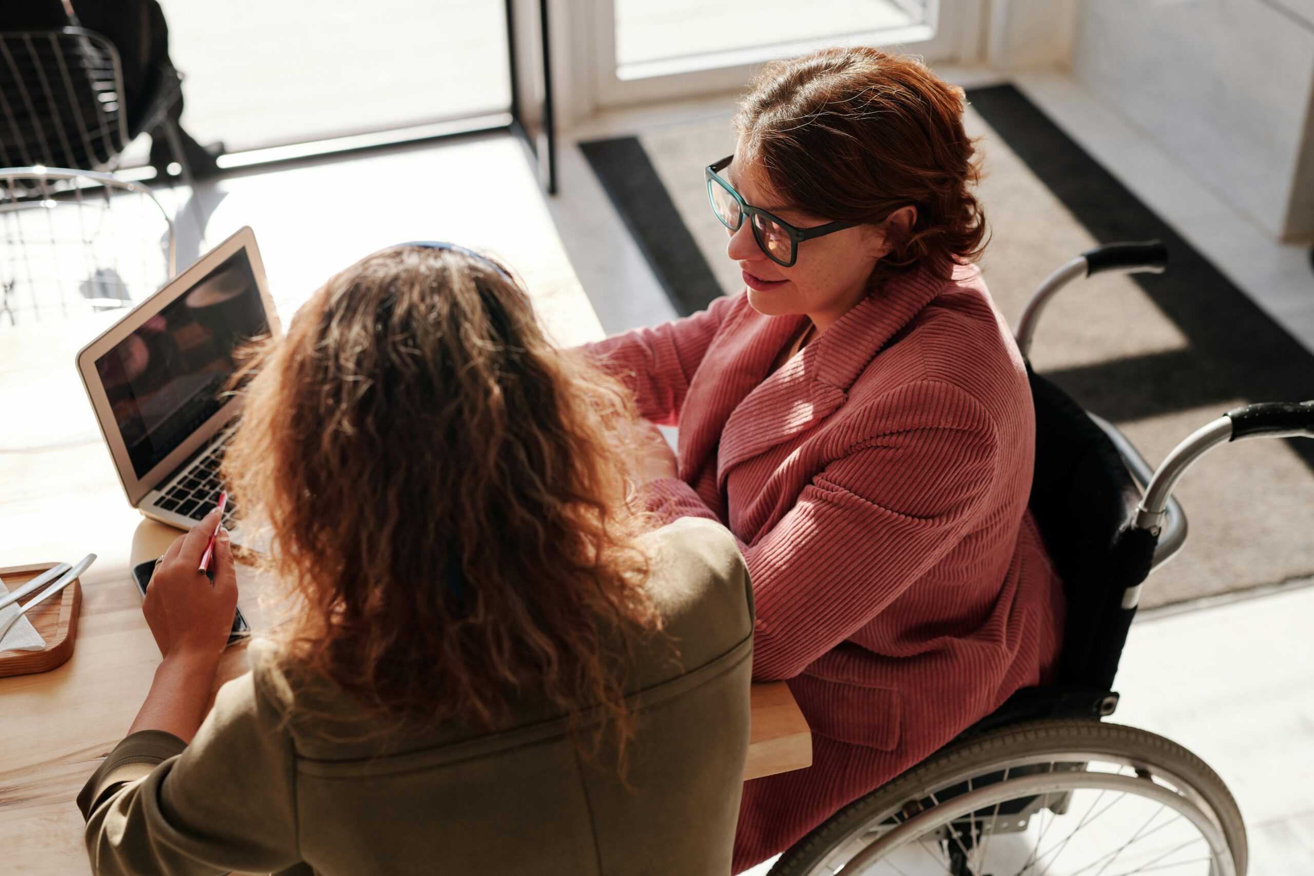 A woman in a wheelchair discusses work with a colleague at a desk, using a laptop in a sunlit office or cafe setting.