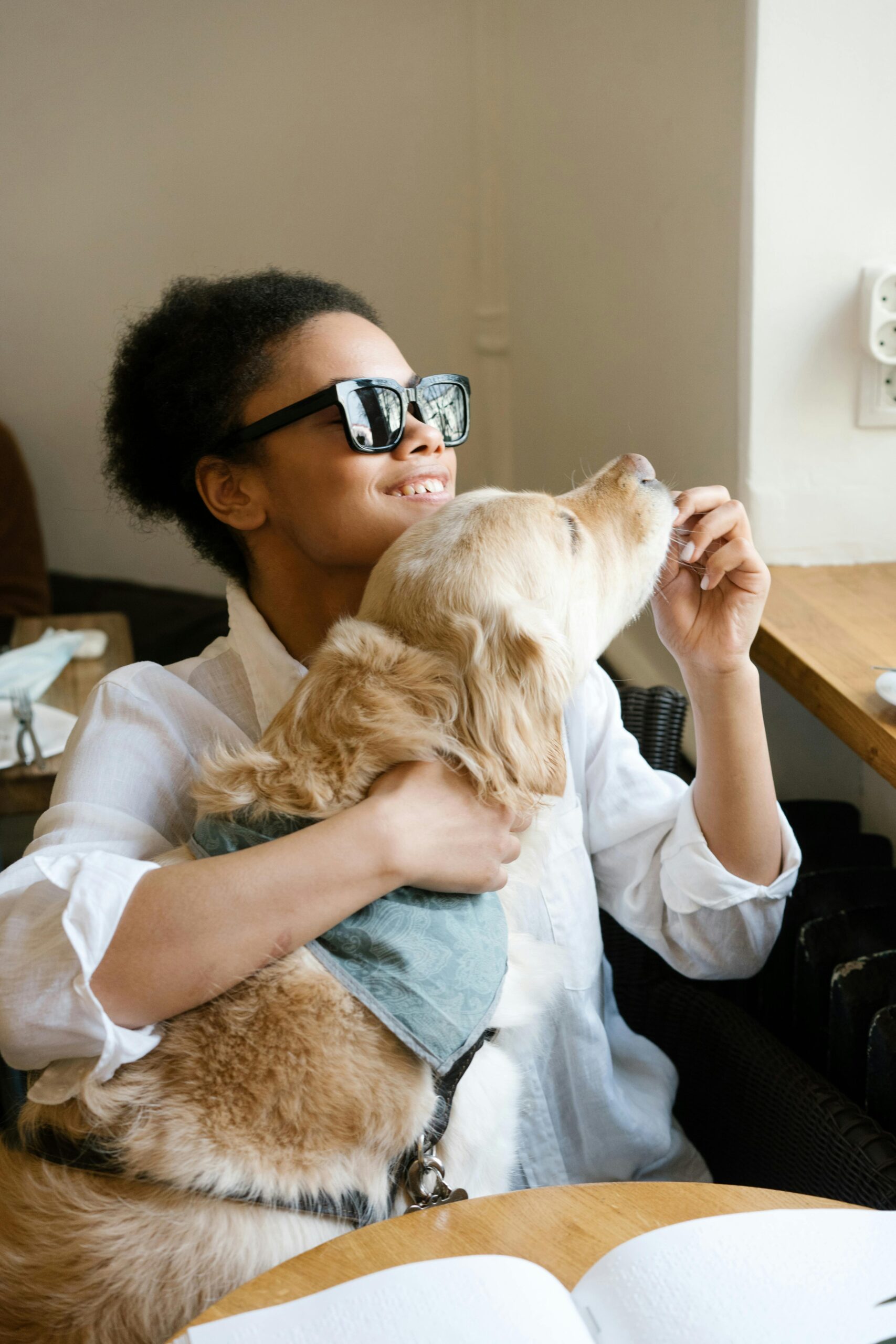 A smiling woman wearing dark sunglasses holds a golden retriever close while offering it a treat, seated indoors at a wooden table. The scene conveys warmth and companionship, suggesting support for individuals with visual impairments and service animals.