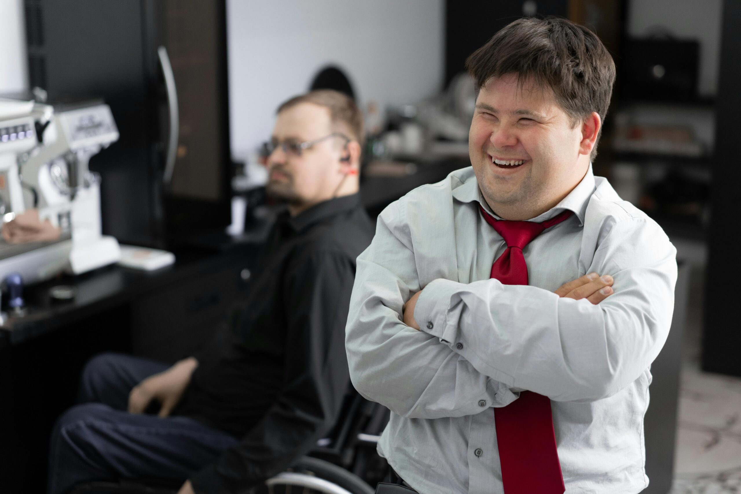 A cheerful man with Down syndrome wearing a white shirt and red tie stands confidently with arms crossed in the foreground. In the background, a man using a wheelchair appears focused in a professional setting. The image promotes workplace inclusion and diversity.