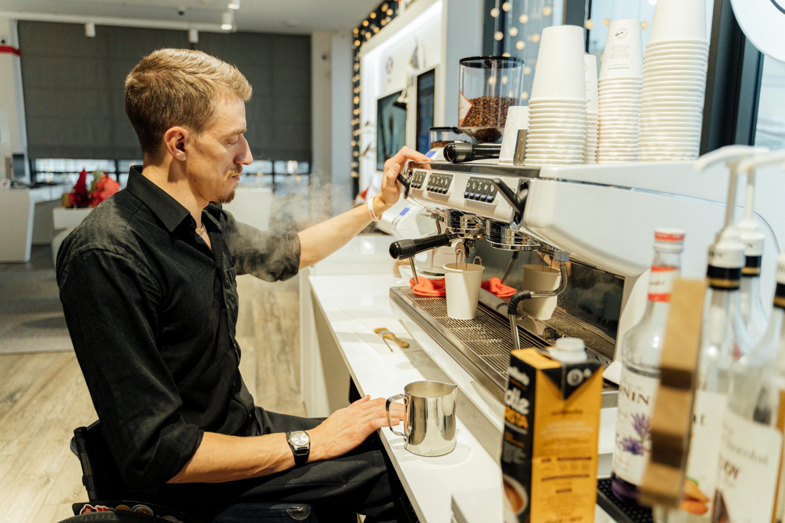 A male barista using a wheelchair prepares coffee at an espresso machine in a modern café.